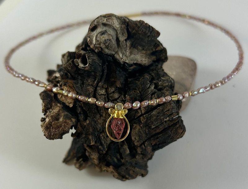 Kette aus kleinen rosafarbenen Perlen mit goldenen Zwischenstücken; als Anhänger ein roter Stein in einem tropfenförmigen Goldrahmen, darüber ein kleiner heller Stein