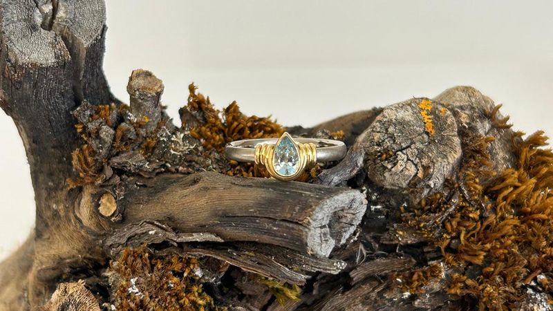 Ring mit hellem Band und einem tropfenförmigen, hellblauen Stein in goldener Fassung, auf einem Ast mit Moos und Flechten
